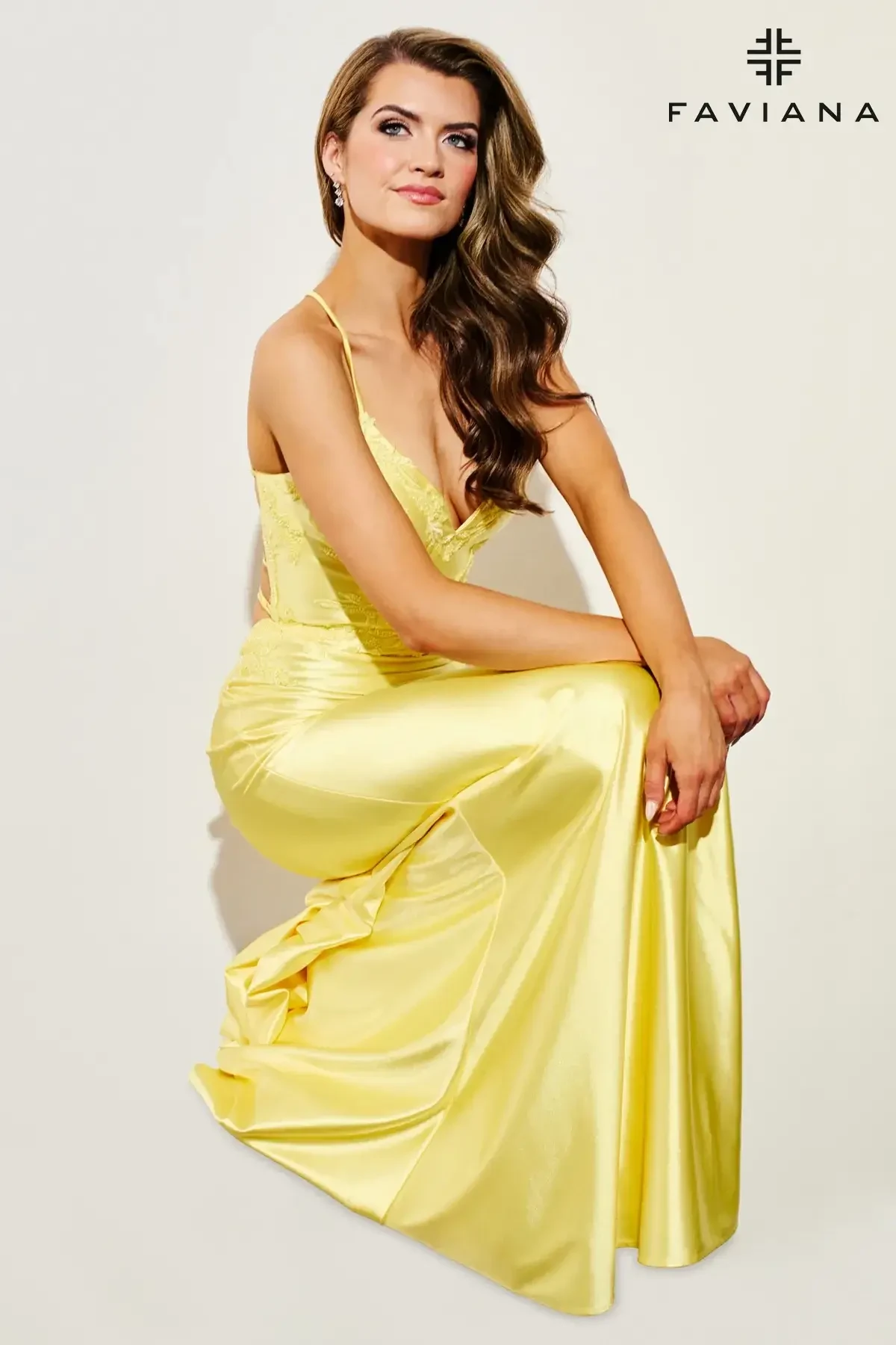 A woman in a flowing yellow gown kneels gracefully against a plain background. She looks confidently ahead, with soft waves in her hair, conveying elegance.