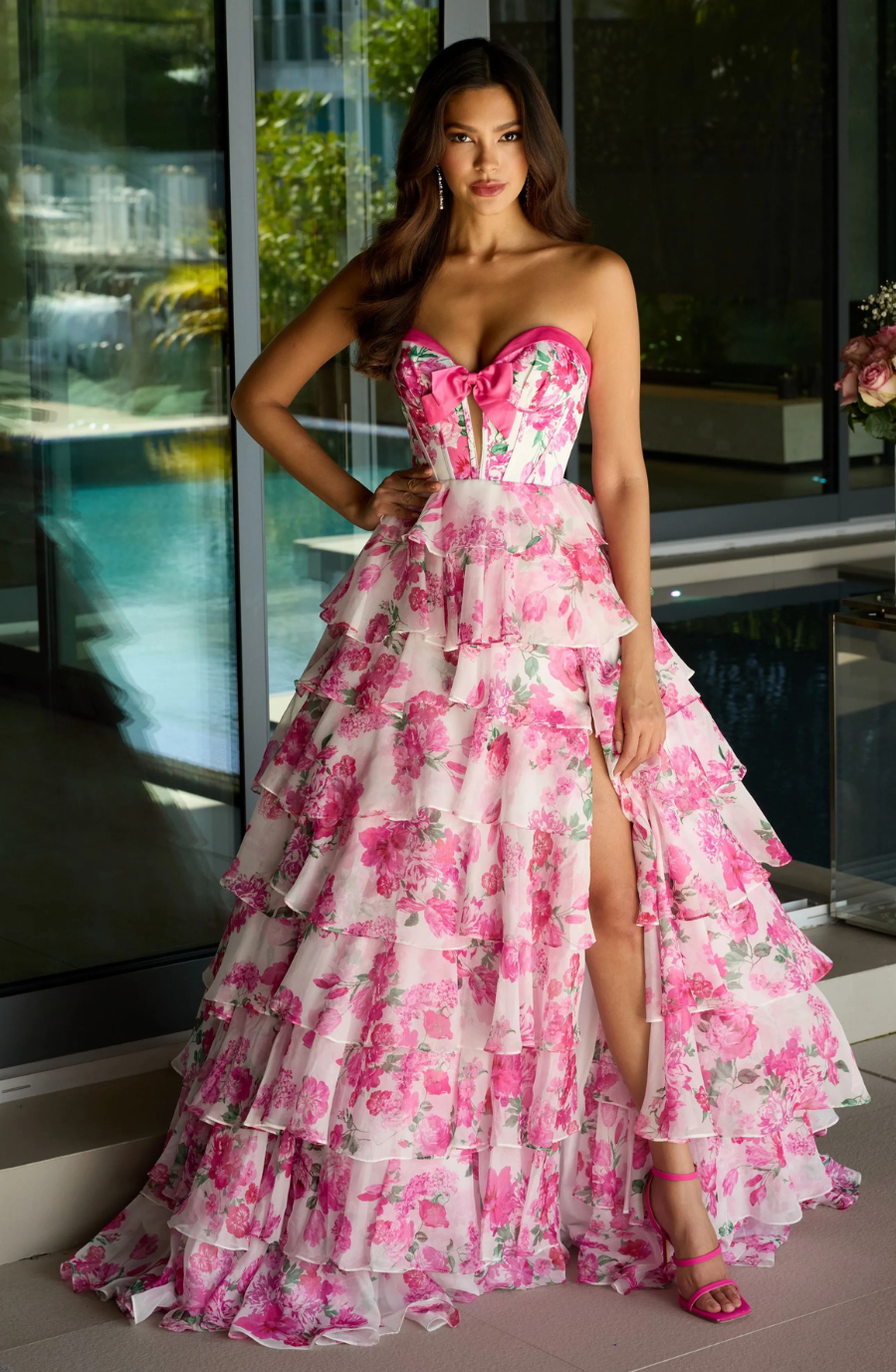 A woman in a floral pink and white ruffled gown poses confidently near large windows. The dress has a thigh-high slit, complemented by matching pink heels. The setting is bright and elegant, with a serene pool visible outside.