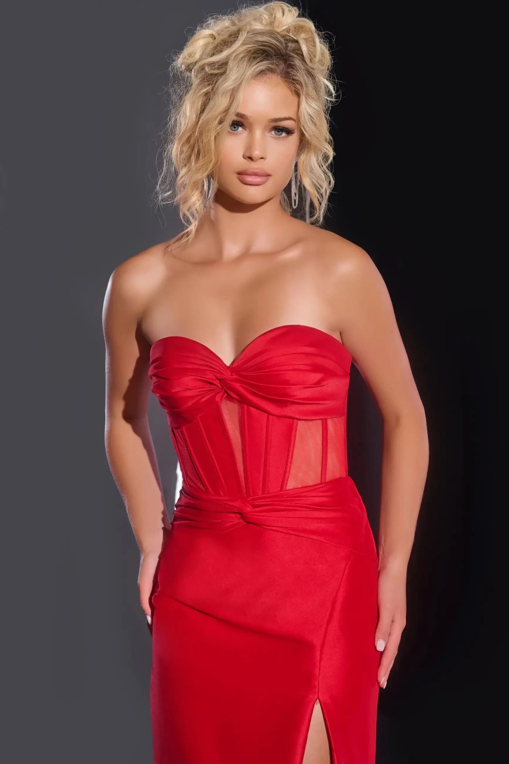Model is wearing a strapless red fitted prom dress with a corset bodice and thigh high slit