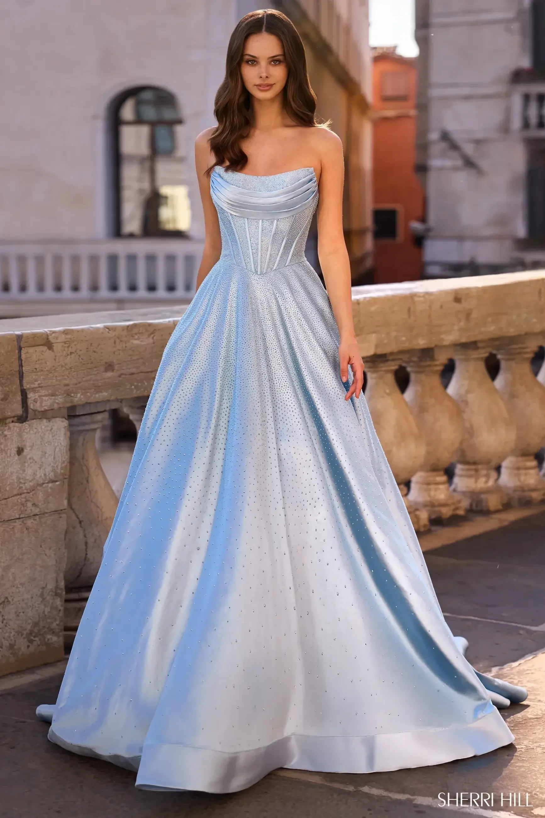 A model wearing a light blue strapless gown stands on a stone balcony, with buildings in the background. The gown has a fitted bodice and flows into a full skirt.
