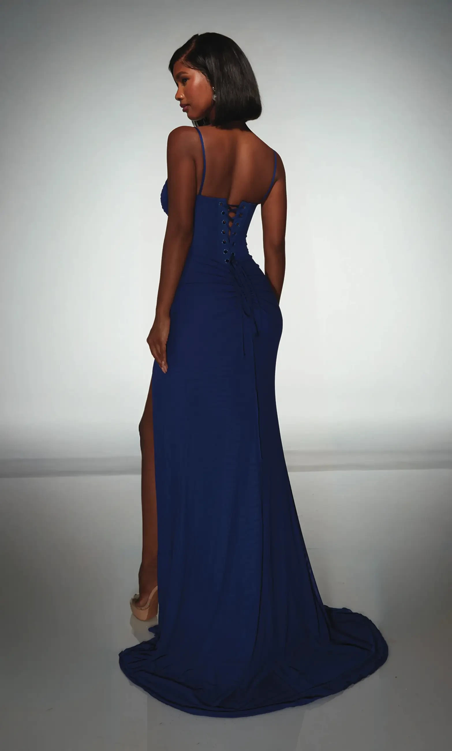 A woman wearing a blue gown, viewed from the back. The dress has a thigh-high slit and spaghetti straps.